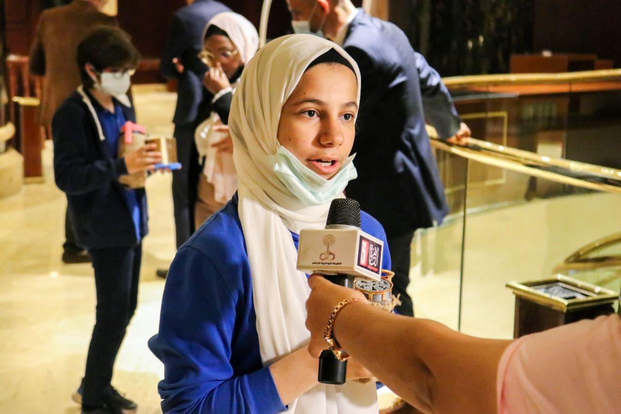 A young person with a hijab being interviewed at IVY STEM International School. They are holding a microphone with a logo, indicating their participation in a media interview or public speaking event. Other individuals in the background can be seen using smartphones and adjusting face masks. The setting suggests an indoor venue, possibly a hotel lobby or conference center, with formal decor and lighting.